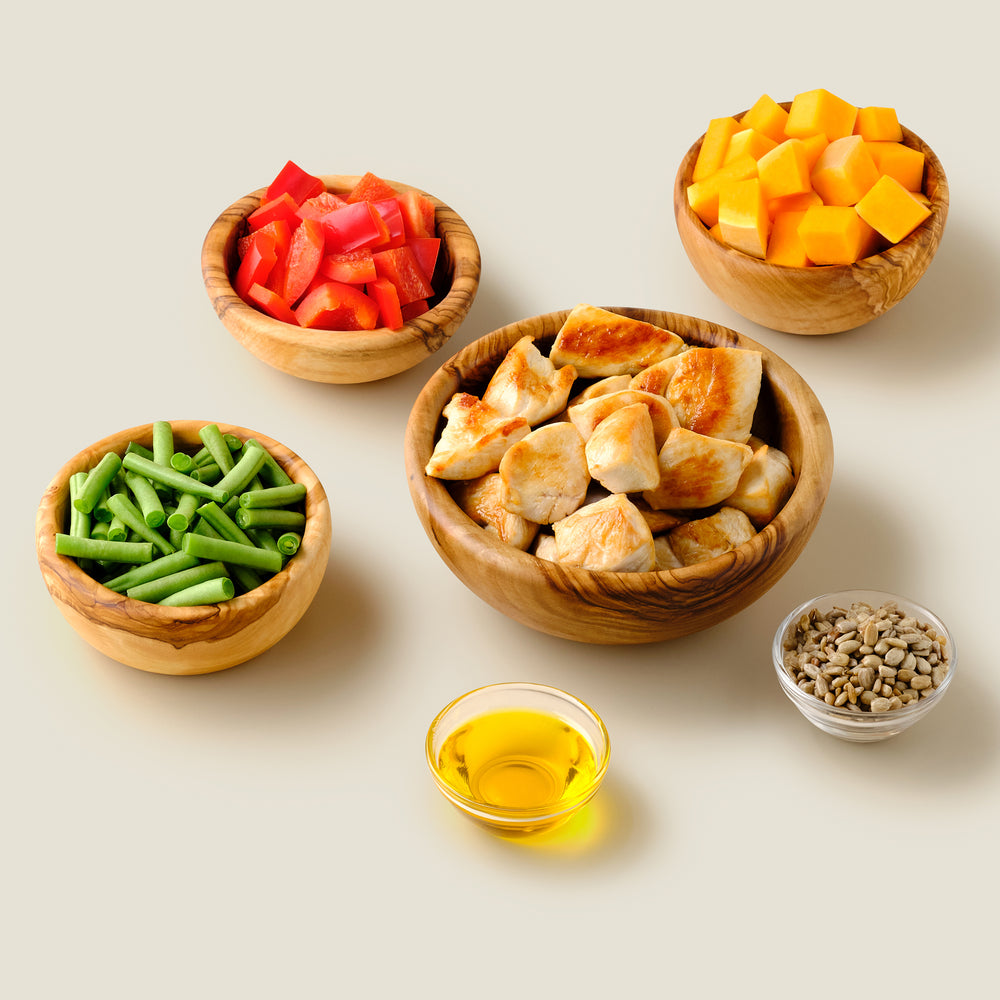 Fresh ingredients for organic grain-free chicken dog food, including organic chicken and vegetables, displayed in wooden bowls.